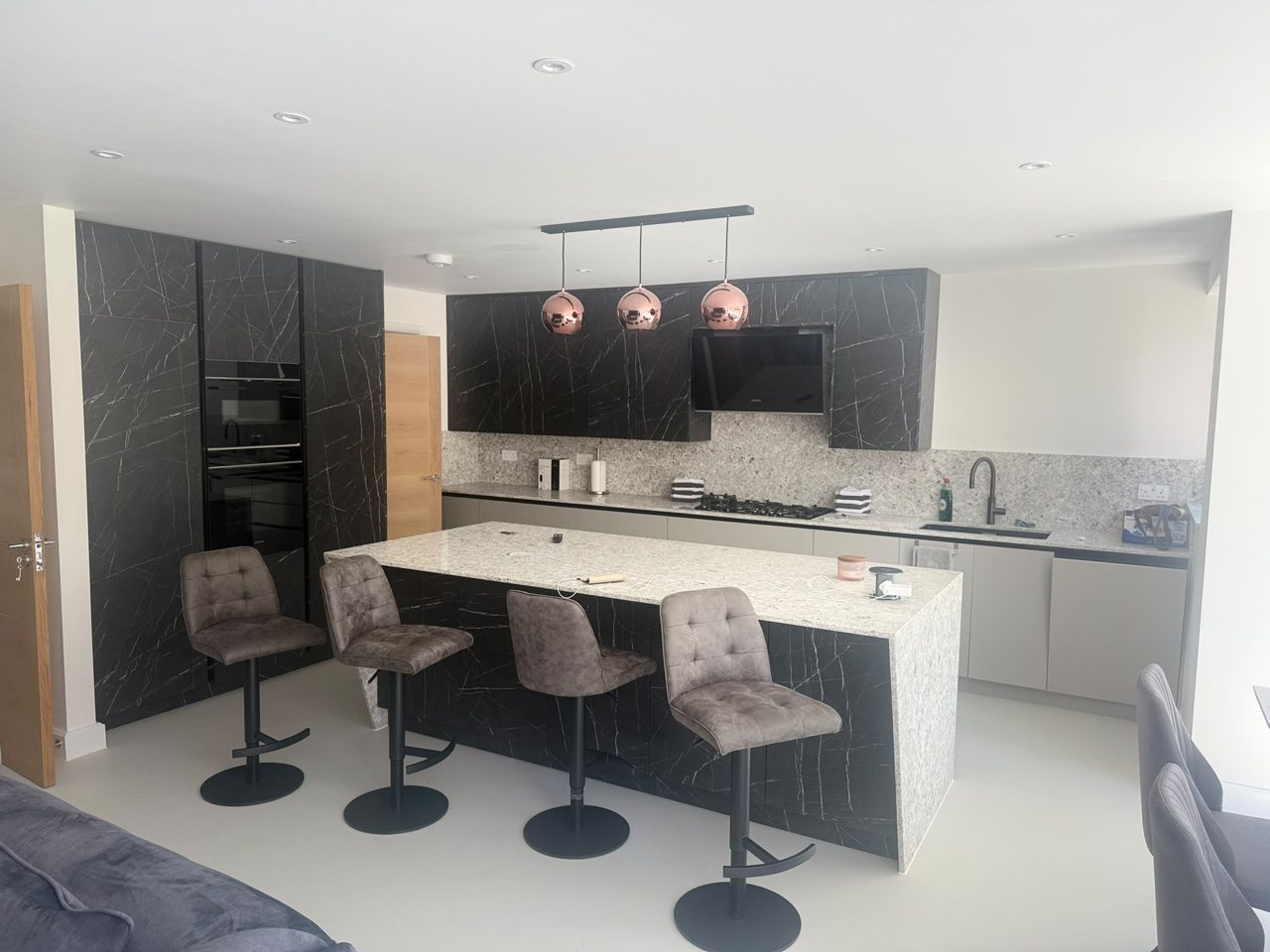 Completed MALCON contemporary kitchen with a large central island, black marble-effect cabinetry, a light stone island with bar seating and copper pendant lighting